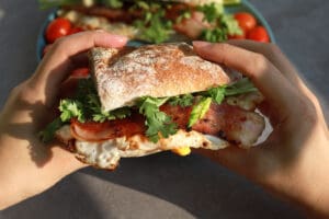 Hand holding a fresh baguette sandwich with bacon, fried egg, parsley and grilled cherry tomatoes.